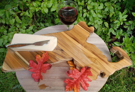 Massachusetts state-shaped teak wood charcuterie board with natural wood grain, shown with cheese and wine for scale.
